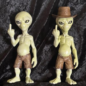Può includere: Due statuette di alieni verde pallido con grandi occhi scuri, ognuna indossa pantaloncini marroni e fa il gesto del dito medio. Un alieno è a testa scoperta, mentre l'altro indossa un cappello da cowboy marrone con un motivo in rilievo.