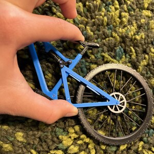 Puede incluir: Un peque&ntilde;o modelo de bicicleta de monta&ntilde;a azul con neum&aacute;ticos negros y un cuadro detallado. La bicicleta en miniatura est&aacute; sujeta por una mano, mostrando su intrincado dise&ntilde;o y sus caracter&iacute;sticas realistas. Las ruedas tienen un patr&oacute;n de radios detallado.