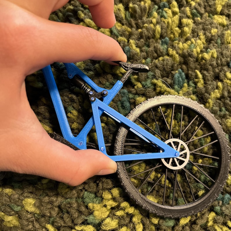 Puede incluir: Un peque&ntilde;o modelo de bicicleta de monta&ntilde;a azul con neum&aacute;ticos negros y un cuadro detallado. La bicicleta en miniatura est&aacute; sujeta por una mano, mostrando su intrincado dise&ntilde;o y sus caracter&iacute;sticas realistas. Las ruedas tienen un patr&oacute;n de radios detallado.