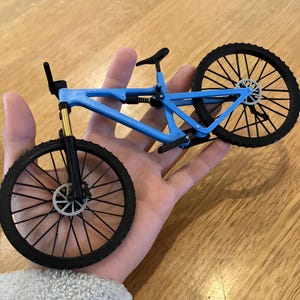 May include: A miniature blue mountain bike with black tires and a black seat. The bike has gold-colored front suspension and a detailed frame. The wheels have intricate spoke designs, and the bike is held in a hand.