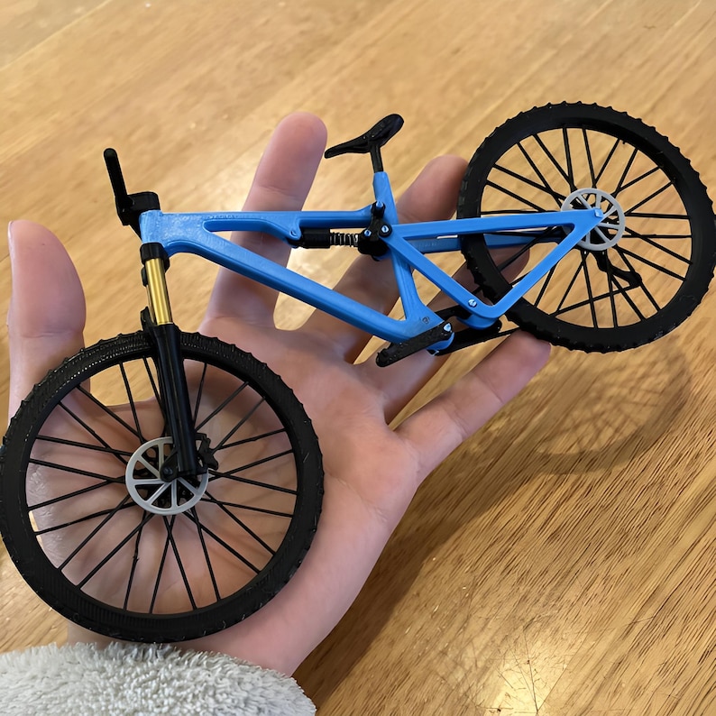 Puede incluir: Una bicicleta de monta&ntilde;a azul en miniatura con neum&aacute;ticos negros y un asiento negro. La bicicleta tiene suspensi&oacute;n delantera dorada y un cuadro detallado. Las ruedas tienen dise&ntilde;os de radios intrincados, y la bicicleta se sostiene en una mano.