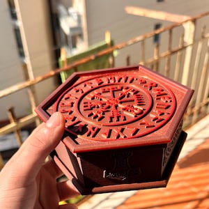 Puede incluir: Una caja hexagonal de color marrón oscuro con una tapa que presenta un diseño circular rojo en relieve. La tapa muestra un símbolo Vegvisir central rodeado por un anillo de caracteres rúnicos. El lateral tiene un relieve de un martillo Mjolnir.