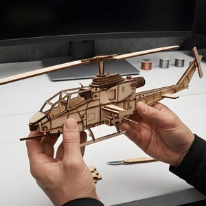 May include: A light brown wooden model helicopter featuring a detailed cockpit, a main rotor blade, and a tail rotor. The structure is composed of laser-cut wooden pieces assembled to form the fuselage, landing skids, and side-mounted weapon pods.