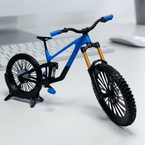 May include: A miniature mountain bike model featuring a blue and black frame, black handlebars with blue grips, gold-coloured front suspension forks, and black knobby tyres. The bicycle is supported by a black stand attached to the rear wheel.