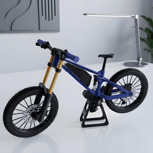 May include: A miniature blue mountain bike model featuring gold-coloured front suspension forks, black knobby tyres, a rear shock absorber, and a disc brake system. The bicycle is mounted on a small black stand, showcasing a detailed frame design.