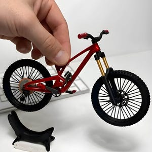 May include: A miniature red mountain bike model featuring black spoked wheels, gold-coloured front suspension forks, and red handlebar grips. The frame is metallic red with a detailed drivetrain and a small black display stand with the text SCALE-DOWN_01.