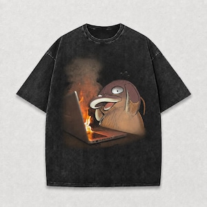 May include: Black, oversized t-shirt featuring a cartoon illustration of a brown and tan creature with a laptop on fire. The creature has a surprised expression. The shirt has a washed, vintage look.