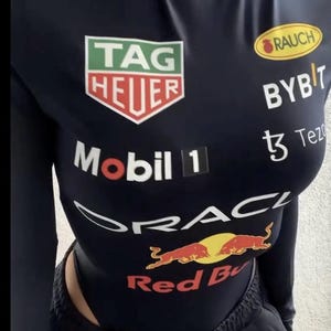 May include: Navy blue bodysuit with multiple sponsor logos, including "TAG HEUER", "Mobil 1", and "Red Bull". The long-sleeved top has a mock neck and is paired with black sweatpants. The overall design is sporty and features a racing theme.