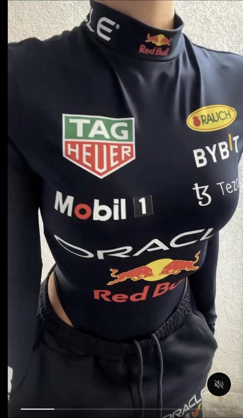 May include: Navy blue bodysuit with multiple sponsor logos, including "TAG HEUER", "Mobil 1", and "Red Bull". The long-sleeved top has a mock neck and is paired with black sweatpants. The overall design is sporty and features a racing theme.