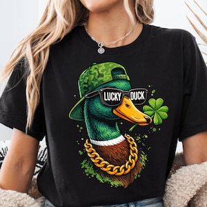 Puede incluir: Camiseta negra con un diseño de pato de dibujos animados. El pato lleva gafas de sol, una gorra de camuflaje y una cadena dorada. Sostiene un trébol de cuatro hojas y las gafas de sol dicen "LUCKY DUCK". Un diseño divertido con temática del Día de San Patricio.