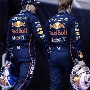 May include: Two people wearing navy blue racing suits with orange accents and the Red Bull logo on the back. One holds a helmet with a white, blue, and red design. The other wears a navy blue cap with a white number 1.
