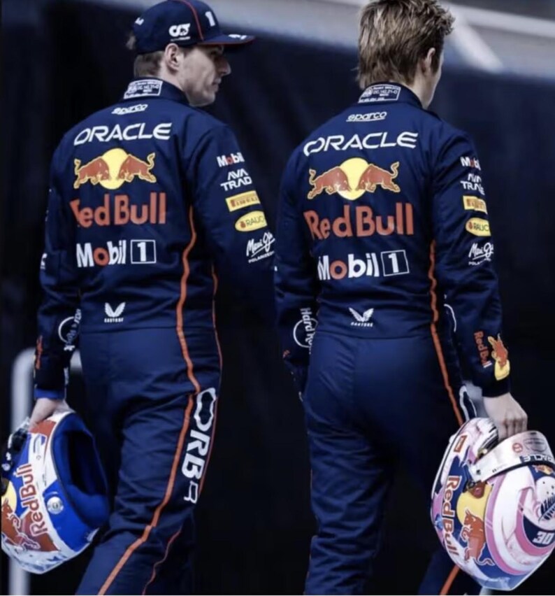 May include: Two people wearing navy blue racing suits with orange accents and the Red Bull logo on the back. One holds a helmet with a white, blue, and red design. The other wears a navy blue cap with a white number 1.