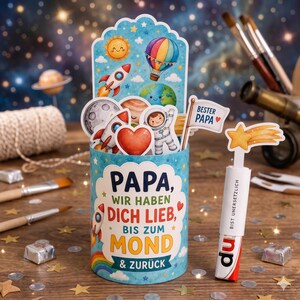 Puede incluir: Un recipiente cilíndrico azul decorado con ilustraciones espaciales que incluyen cohetes, planetas y un sol. El texto dice: "PAPA, WIR HABEN DICH LIEB, BIS ZUM MOND & ZURÜCK." Una pequeña bandera blanca adjunta muestra "BESTER PAPA" con un corazón.
