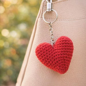 Op de afbeelding: Een rode gehaakte hartvormige sleutelhanger die aan een zilveren ketting hangt die aan een lichtroze handtas is bevestigd. Het hart is gemaakt van getextureerd garen en heeft de vorm van een hart. De handtas heeft een rits en een zilveren ring.