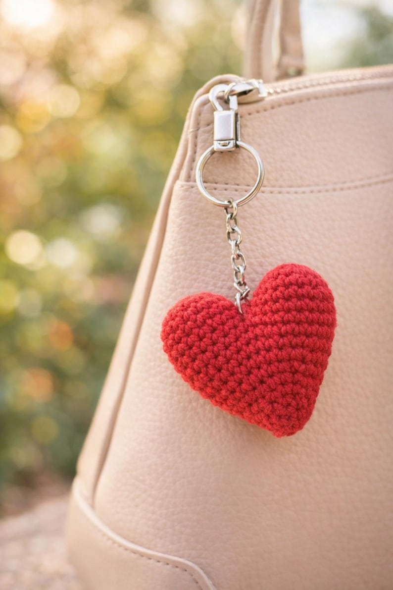Op de afbeelding: Een rode gehaakte hartvormige sleutelhanger die aan een zilveren ketting hangt die aan een lichtroze handtas is bevestigd. Het hart is gemaakt van getextureerd garen en heeft de vorm van een hart. De handtas heeft een rits en een zilveren ring.