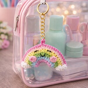 Op de afbeelding: Een kleurrijke, gehaakte regenboogsleutelhanger met een lachend gezicht, bevestigd aan een gouden ketting en ring. De regenboog heeft roze, gele, groene en blauwe tinten. De sleutelhanger hangt voor een doorzichtige roze cosmetische tas.