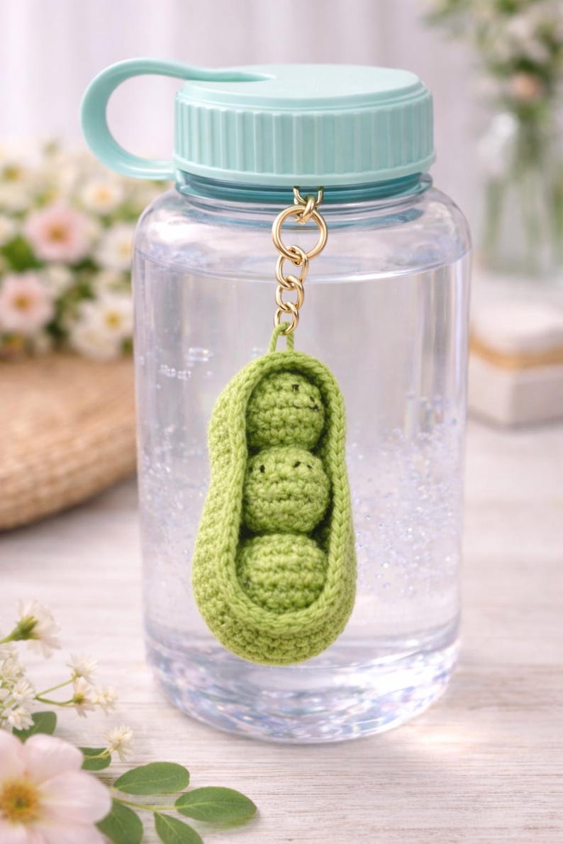Op de afbeelding: Een doorzichtige waterfles met een lichtblauwe dop en handvat, gevuld met water. Een groene gehaakte erwtenscheut-bedel met drie erwten hangt aan de dop.