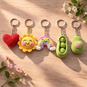 May include: A collection of five crochet keychains. The keychains include a red heart, a smiling yellow sun, a rainbow, a pea pod with three peas, and an avocado. Each keychain has a silver ring and chain.