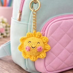 Op de afbeelding: Een gele gehaakte zonnesleutelhanger met een lachend gezicht en roze wangen hangt aan een gouden ketting die is bevestigd aan een lichtblauwe en roze rugzak. De zon heeft een gedetailleerd, getextureerd uiterlijk en de rugzak heeft een gewatteerd patroon.