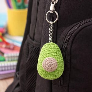Op de afbeelding: Een groene gehaakte avocado sleutelhanger met een bruine pit hangt aan een zilveren ketting die is bevestigd aan een zwarte rugzakrits. De avocado is het middelpunt, met een gedetailleerd, gestructureerd uiterlijk. De achtergrond bevat een gele beker en kleurpotloden.