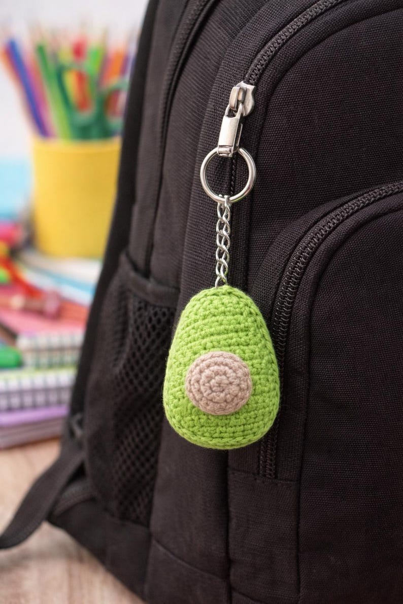 Op de afbeelding: Een groene gehaakte avocado sleutelhanger met een bruine pit hangt aan een zilveren ketting die is bevestigd aan een zwarte rugzakrits. De avocado is het middelpunt, met een gedetailleerd, gestructureerd uiterlijk. De achtergrond bevat een gele beker en kleurpotloden.
