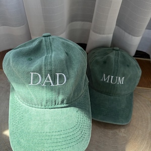 Puede incluir: Dos gorras de béisbol de color verde salvia con una textura suave y cepillada. La parte delantera de la primera gorra presenta la palabra DAD bordada en hilo blanco, mientras que la segunda gorra detrás tiene la palabra MUM bordada en blanco.