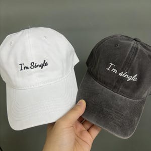 Può includere: Sono mostrati due cappellini da baseball, uno bianco e uno grigio antracite. Entrambi i cappellini presentano la frase "I'm Single" ricamata in bianco. Il cappellino bianco è tenuto in mano, evidenziando il design.