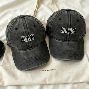 May include: Two charcoal grey baseball caps with a distressed finish. One cap features the white embroidered text DAD, and the other features the white embroidered text MUM. Both caps have a curved brim and sit on a cream-coloured fabric bag.
