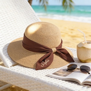 May include: A woven straw sun hat with a wide brim and a dark brown ribbon tied in a bow. The ribbon has the name "Sofia" embroidered on it. Sunglasses and a magazine rest on a white chair near a coconut.