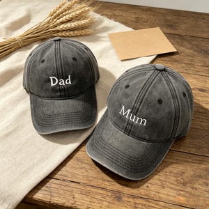 May include: Two grey baseball caps with curved peaks. One cap has the word "Dad" embroidered in white, and the other has "Mum". The caps are made of a worn fabric and are displayed on a wooden surface.