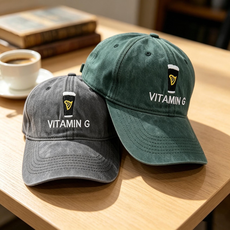 May include: Two baseball caps, one grey and one dark green, are displayed on a wooden surface. Each cap has an embroidered beer glass design and the text "VITAMIN G" in white. The caps are casual and stylish.