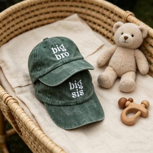 Puede incluir: Dos gorras de béisbol verdes apiladas, una con "big bro" bordado en blanco y la otra con "big sis". Un oso de peluche beige y un sonajero de madera también están en la imagen.