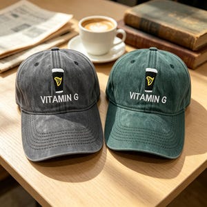 May include: Two baseball caps, one dark gray and one green, sit on a wooden surface. Each cap features an embroidered design of a pint glass with a yellow harp and the text "VITAMIN G". A cup of coffee and books are in the background.