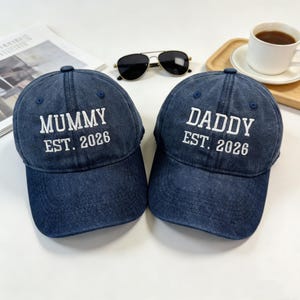 May include: Two navy blue baseball caps with white embroidered text. One cap reads "MUMMY EST. 2026" and the other reads "DADDY EST. 2026". The caps are on a white surface, with sunglasses and a cup of coffee in the background.
