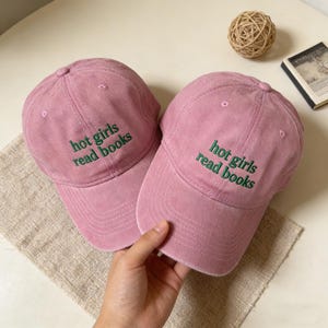 May include: Two pink baseball caps with the embroidered text "hot girls read books" in green. The caps are made of a soft, worn fabric and have a classic six-panel design with a curved brim. A hand is holding one of the caps.