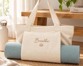 Könnte beinhalten: Eine cremefarbene Canvas-Tragetasche mit zwei stabilen Henkeln und einem äußeren Riemen zur Befestigung einer zusammengerollten hellblauen Yogamatte. Auf der Vorderseite ist das Wort Breathe in brauner Schrift über einem Lotus-Symbol eingestickt.