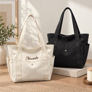 Może przedstawiać: Dwie torby typu tote z płótna, jedna kremowa i jedna czarna, każda z dwoma uchwytami, kieszeniami bocznymi i przednią kieszenią zapinaną na pojedynczy metalowy zatrzask. Na kremowej torbie widnieje wyhaftowane brązowym pismem słowo Necade.