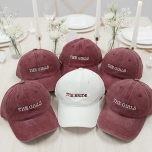 以下が含まれることがあります： 「THE BRIDE」とバーガンディ色で刺繍された白いキャップを中心に、白い「THE GIRLS」の文字が入ったバーガンディ色のキャップが並んだ野球帽のコレクション。花柄の装飾が施されたテーブルにキャップが配置されています。