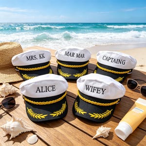 Op de afbeelding: Vijf witte kapiteinshoeden met zwarte randen en gouden accenten liggen op een houten oppervlak. Elke hoed heeft een andere naam in het zwart geborduurd: "MRS. KK", "MAR MAR", "CAPTAIN", "ALICE" en "WIFEY".