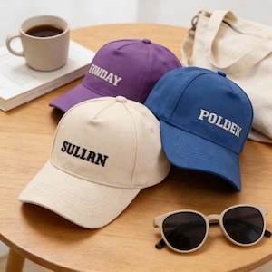 May include: Three baseball caps in beige, purple, and blue, each with white embroidered text. The beige cap reads "SULLAN", the purple cap reads "FONDAY", and the blue cap reads "POLDEK". A pair of sunglasses and a coffee mug are also visible.