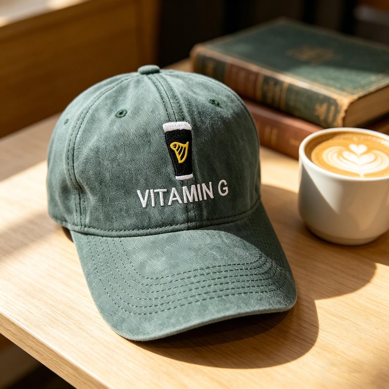 May include: Green baseball cap with an embroidered design of a pint of beer and the text "VITAMIN G" in white. The cap is made of a soft, worn fabric and has a curved brim. A cup of coffee sits next to the cap.