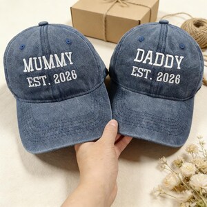 May include: Two blue denim baseball caps with white embroidered text. One cap reads "MUMMY EST. 2026", the other reads "DADDY EST. 2026". The caps have a curved brim and a small button on top. A hand is holding the caps.