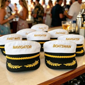 Könnte beinhalten: Weiße Matrosenmützen mit schwarzen Krempen und goldener Stickerei. Die Mützen tragen nautische Bezeichnungen wie "Navigator" und "First Mate". Die Mützen sind auf einer weißen Oberfläche angeordnet, mit einem verschwommenen Hintergrund von Menschen.