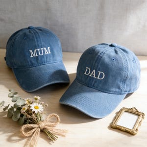 May include: Two blue denim baseball caps, one embroidered with "MUM" and the other with "DAD" in white. A small bouquet of flowers and a decorative gold frame are in the foreground.