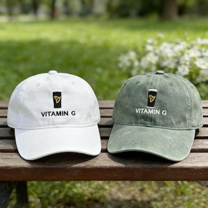 Könnte beinhalten: Zwei Baseballkappen, eine weiße und eine olivgrüne, liegen nebeneinander auf einer Holzbank. Jede Kappe hat den gestickten Text "VITAMIN G" unter einer kleinen Grafik in Schwarz und Gold.