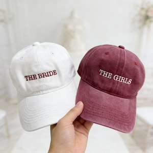May include: Two baseball caps are shown, one white and one burgundy. The white cap features "THE BRIDE" embroidered in burgundy, and the burgundy cap says "THE GIRLS" in white. Both caps have a curved peak.