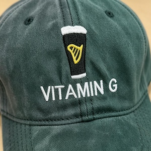 May include: A dark green baseball cap with a black and yellow beer glass design. The text "VITAMIN G" is embroidered in white below the design. The cap has a curved brim and a six-panel construction.