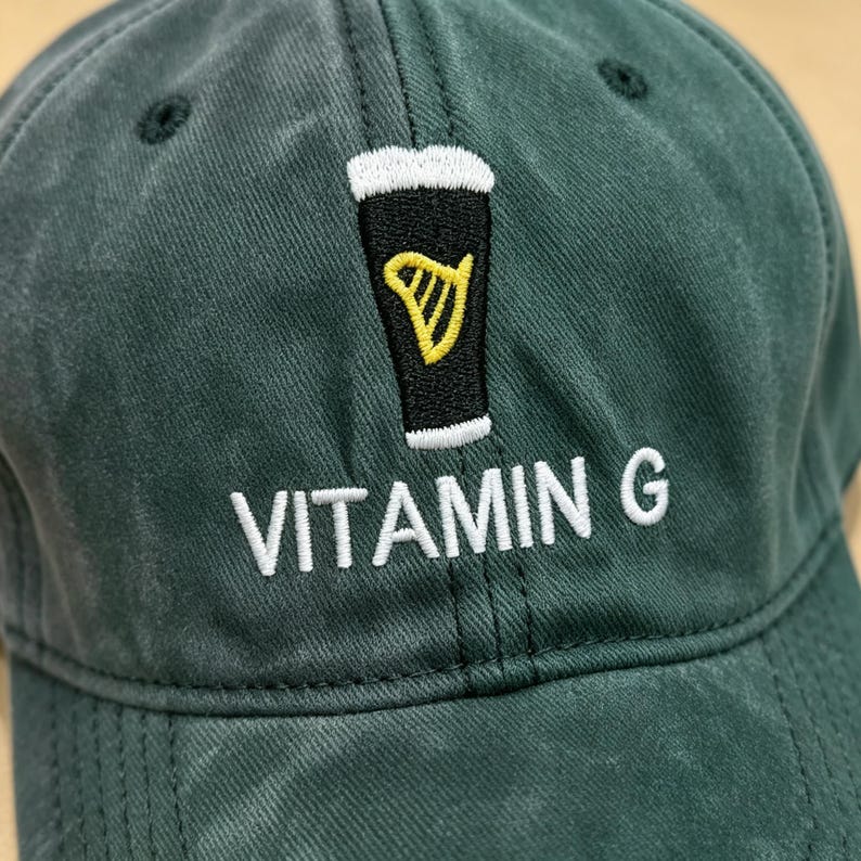 May include: A dark green baseball cap with a black and yellow beer glass design. The text "VITAMIN G" is embroidered in white below the design. The cap has a curved brim and a six-panel construction.
