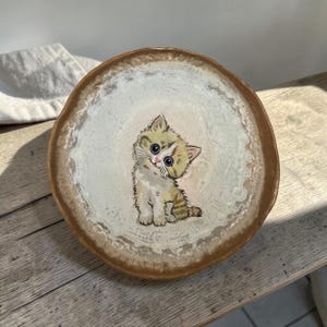 Puede incluir: Un plato de cerámica redondo con un borde marrón irregular y texturizado y un centro de color crema. El plato presenta una ilustración pintada a mano de un gatito pequeño y esponjoso con ojos oscuros grandes, pelaje marrón claro y marcas blancas.