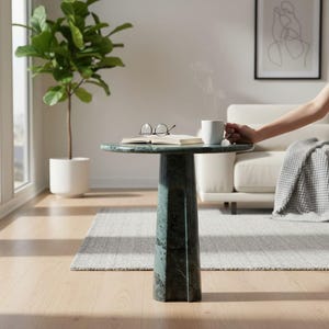 Puede incluir: Una mesa auxiliar de mármol verde con una parte superior redonda y una base geométrica. Un libro y gafas descansan sobre la mesa, y una taza blanca con vapor es sostenida por una persona. Una planta en maceta blanca está en el fondo.